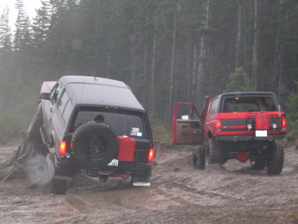 Red 1987 Ford Bronco with open driver door and black Bronco with camper shell on muddy terrain