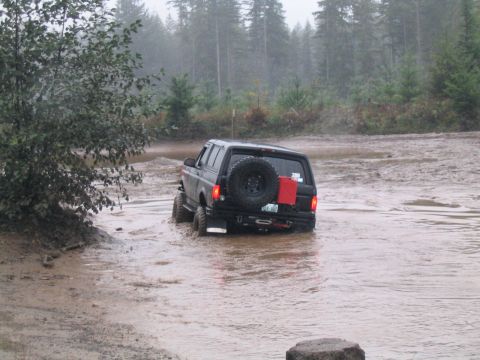 1989 Ford Bronco - A black 1989 Ford Bronco navigates through a muddy water crossing in a wooded area.