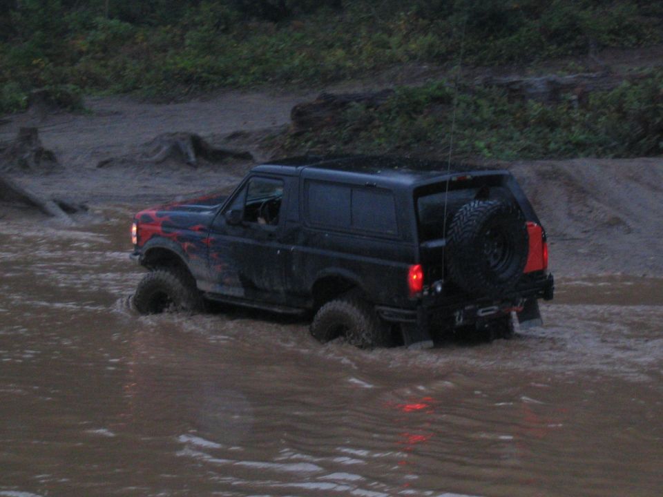 Black 1989 Ford Bronco with red flame decals driving through muddy water