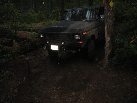 1989 Ford Bronco - A dark-colored SUV is seen on a muddy forest trail, partially obscured by trees.