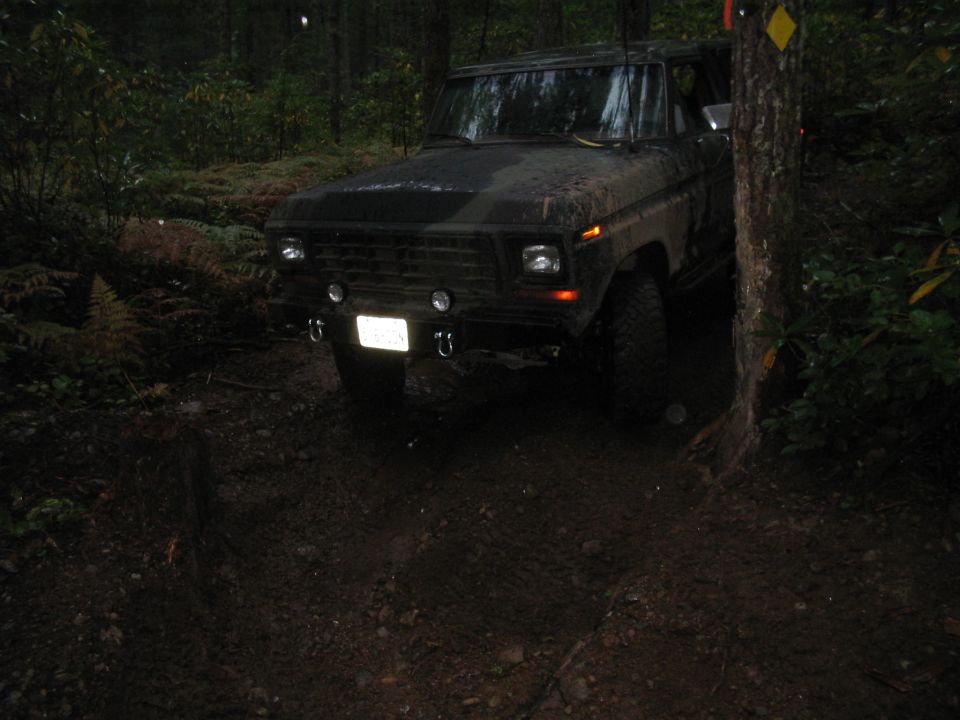 Dark-colored SUV with a rectangular grille and square headlights, parked on a muddy trail in a forest