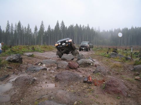 1989 Ford Bronco - Two vehicles are shown off-roading on a challenging, wet, and rocky course with a dense forest in the background.