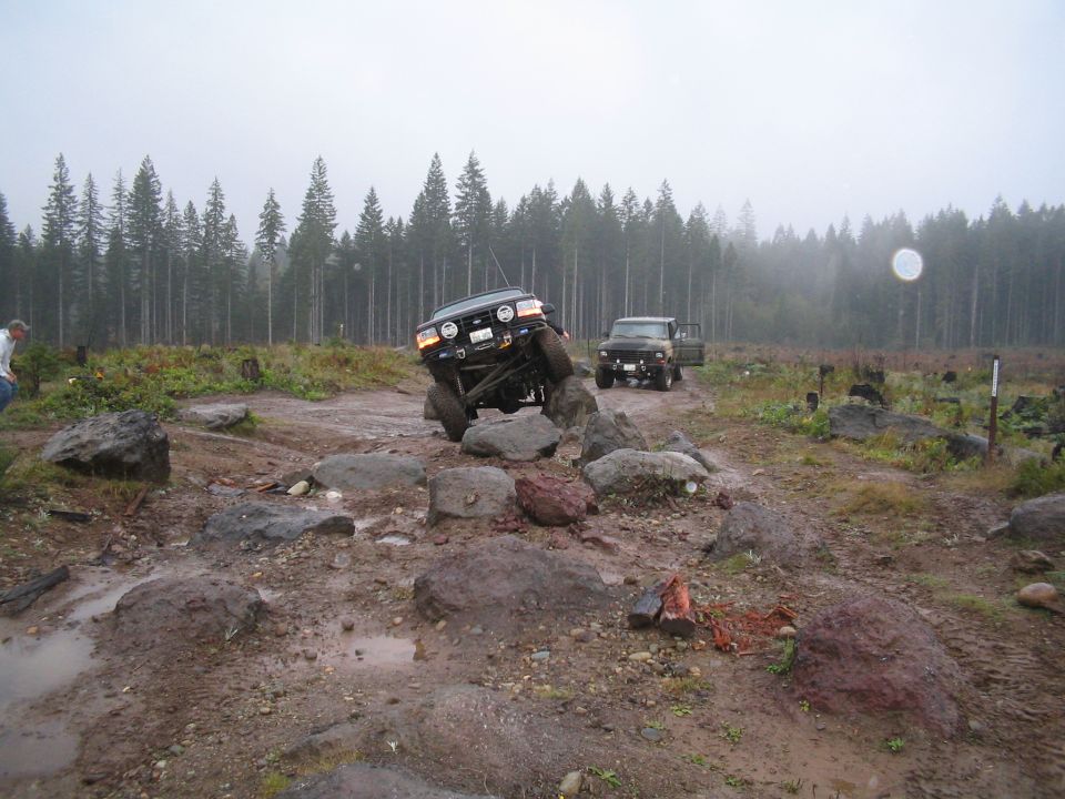 Two off-road vehicles navigating a rocky, muddy trail with trees in the background. One vehicle is tilted on rocks