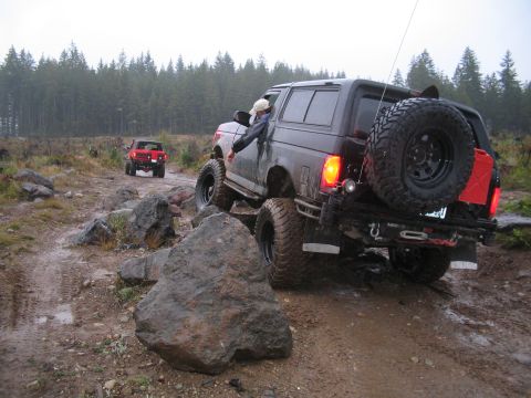 1989 Ford Bronco - A dark SUV drives off-road on a wet, rocky path, with another vehicle following in the distance.