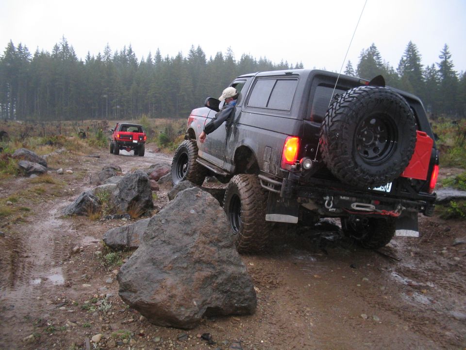 Dark SUV with large tires navigating a rocky, muddy trail, with a red SUV behind it