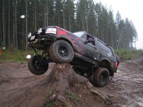 1989 Ford Bronco - A modified black Ford Bronco with flame decals navigates an off-road obstacle on a muddy trail.
