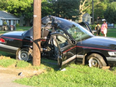 1999 Ford Crown Victoria - A black full-size sedan with a red bodyside pinstripe wrapped around a wooden utility pole after a crash, the driver's side caved in at the…