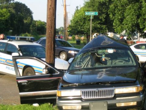 1999 Ford Crown Victoria - A black sedan with a heavily damaged roof sits at an intersection next to a police car.