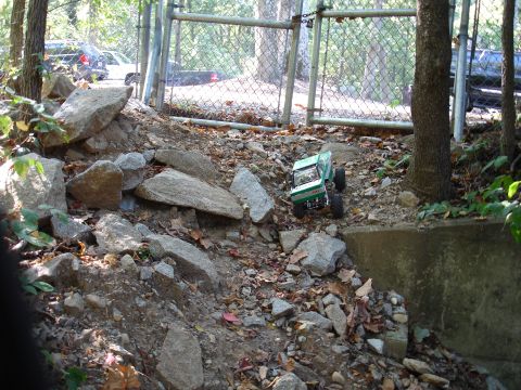 1996 Ford Bronco - A green-and-white radio-controlled rock crawler climbing a rocky leaf-covered embankment beside a chain-link gate and a concrete retaining…