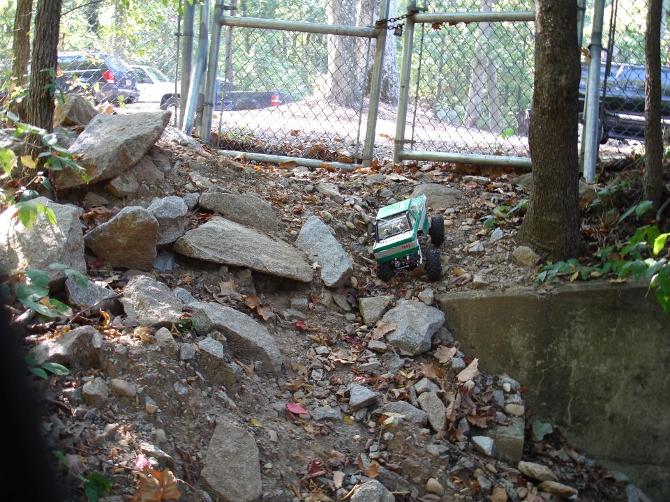 Green and white RC rock crawler climbing a rocky leaf-covered bank by a chain-link gate