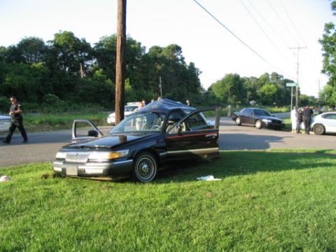 1999 Ford Crown Victoria - Dark Mercury sedan on a grassy roadside with door open, chrome grille and wire wheels; other cars and bystanders behind.