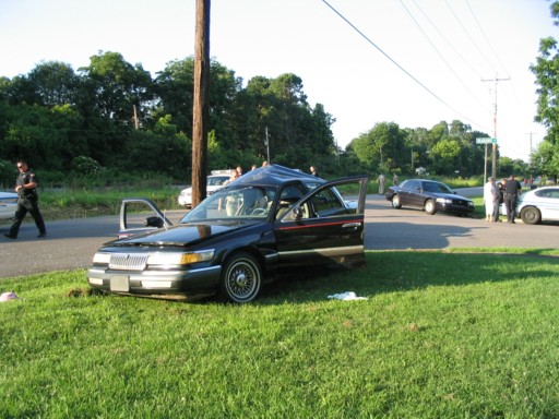 Black Mercury sedan parked on grass roadside with driver door open and people in background