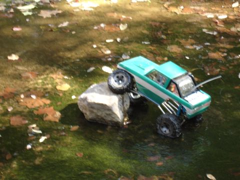 1996 Ford Bronco - Overhead view of a teal-and-white radio-controlled crawler styled like a full-size Bronco, flexed with one rear tire up on a rock in shallo…