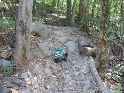 1996 Ford Bronco - A green-and-white radio-controlled rock crawler descending a rocky, leaf-strewn slope in the woods, front wheels dropping over a ledge of f…