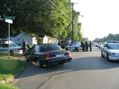 1999 Ford Crown Victoria - Police officers interact near several vehicles on a street corner with a street sign reading 'Adams'.