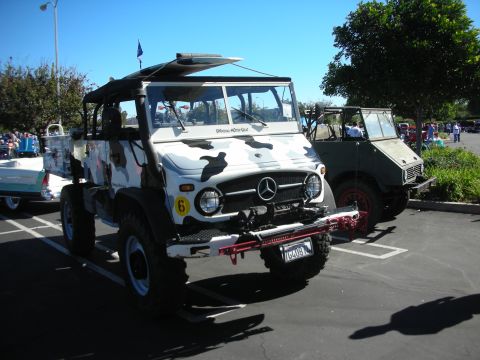 1998 Ford Crown Victoria - white camouflage-painted Mercedes-Benz Unimog 4x4 with a roof rack parked at an outdoor car show.