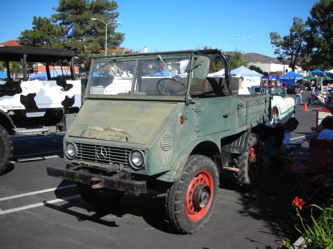1998 Ford Crown Victoria - weathered gray Mercedes-Benz Unimog 4x4 truck with red-painted wheels sits among other vehicles at an outdoor show.