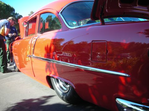 1998 Ford Crown Victoria - Sun-lit side panel of a glossy orange-red lowered classic coupe with chrome side trim, photographed at an outdoor car show.