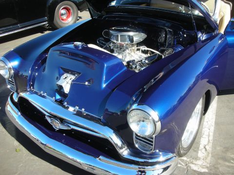 1998 Ford Crown Victoria - deep blue 1950s-style custom coupe with its hood up showing a polished chrome V8 and a smiling face painted under the hood.