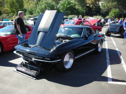 1998 Ford Crown Victoria - black mid-1960s Corvette Sting Ray coupe with its hood raised, displayed with a spec placard at an outdoor car show.