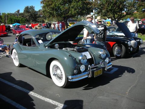 1998 Ford Crown Victoria - grey Jaguar XK140 coupe with whitewall tires and wire wheels sits hood-up beside a black sports car at a crowded outdoor show.
