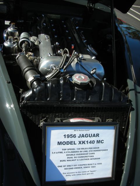 1998 Ford Crown Victoria - polished inline-six engine with dual SU carburetors above a printed placard identifying it as a 1956 Jaguar XK140 MC.