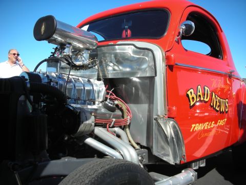 1998 Ford Crown Victoria - red rat-rod pickup with an exposed blown V8 and tall scoop, the cab door lettered Bad News Travels Fast.