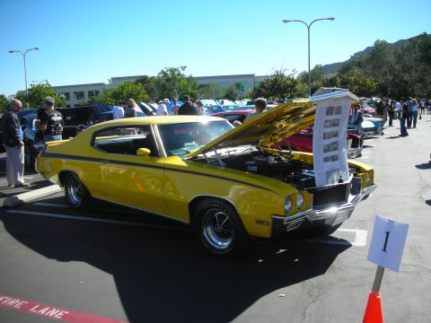 1998 Ford Crown Victoria - yellow muscle car fastback with a black side stripe and open hood at a car show, a placard marked 1 standing beside it.