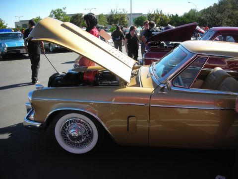 1998 Ford Crown Victoria - gold 1950s Studebaker hardtop coupe with wire wheels and whitewalls, hood raised, among cars at a sunny outdoor show.