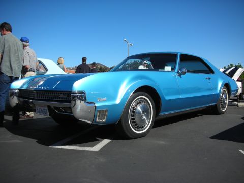 1998 Ford Crown Victoria - light blue Oldsmobile Toronado two-door coupe with concealed headlights and full wheel covers at a sunny outdoor show.