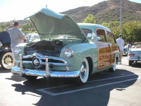1998 Ford Crown Victoria - restored light-green woodie station wagon sits with its hood raised in a sunny car-show parking lot, hills behind.