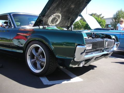 1998 Ford Crown Victoria - Front corner of a dark-green Mercury Cougar with its hood raised, showing the hidden-headlight grille and gray five-spoke wheel.