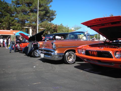 1998 Ford Crown Victoria - copper-brown 1957 Chevrolet sits in a row beside an orange muscle car with its hood up at an outdoor car show.