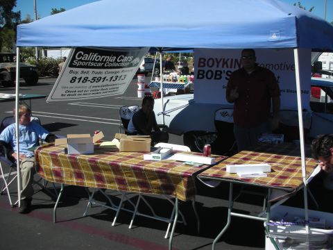 1998 Ford Crown Victoria - People sit under a blue canopy with California Cars and Boykin Bob's Alignment banners and plaid-covered tables.
