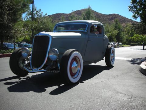1998 Ford Crown Victoria - gray primered 1930s-style hot rod coupe with wide whitewall tires drives through a tree-lined car-show lot.
