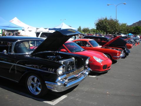 1998 Ford Crown Victoria - black 1957 Chevrolet with hood up leads a row that includes a red Porsche 911 at a busy car show.