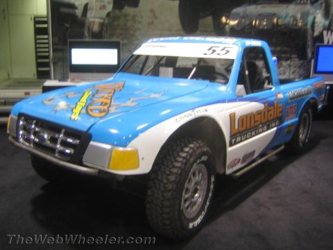 1990 Ford Bronco - blue and white prerunner-style off-road race truck with Lonsdale Trucking sponsorship and number 55 shown at an indoor booth.