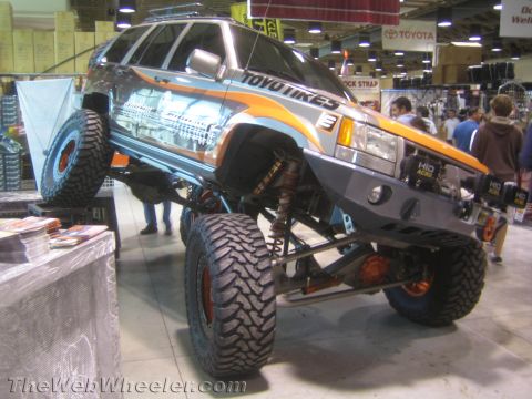 1990 Ford Bronco - Front three-quarter view of a lifted gray-and-orange SUV rock crawler with massive mud tires and Toyo Tires graphics indoors.