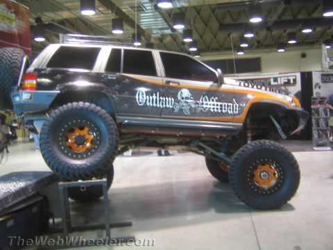 1990 Ford Bronco - Side view of a black lifted SUV rock crawler with 'Outlaw Offroad' graphics, orange beadlock wheels and large mud tires at a show.