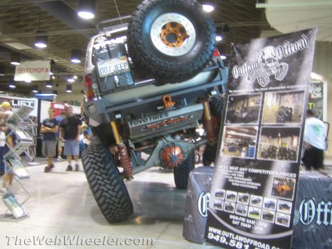 1990 Ford Bronco - lifted off-road SUV displayed next to an Outlaw Offroad promotional banner showing shop photos and a phone number at an indoor show.