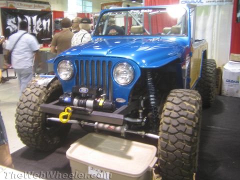 1990 Ford Bronco - blue custom flat-fender Jeep-style 4x4 with round headlights, a slotted grille, front winch and large mud tires on display indoors.