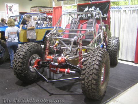 1990 Ford Bronco - polished tube-chassis rock buggy with red coilover shocks, exposed driveline and bucket seats displayed at an indoor expo.