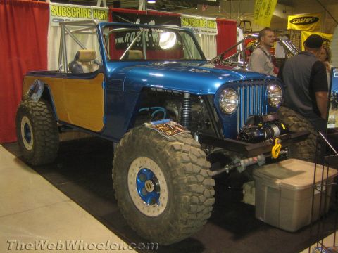 1990 Ford Bronco - Front three-quarter view of a blue custom open-top 4x4 with wood-grain panels, a slotted grille, front winch and oversized tires at a show.
