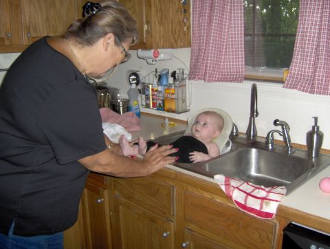 1978 Ford Bronco - A woman bathes a baby in a kitchen sink, with red and white checkered curtains in the background.