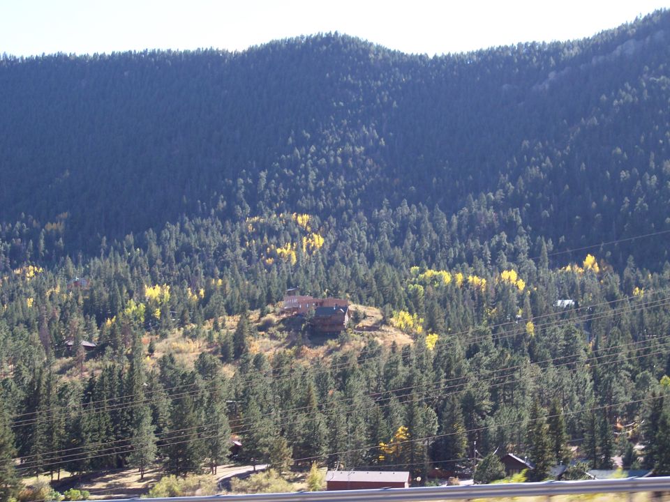 Mountain landscape with dense evergreen trees, some deciduous trees with yellow leaves, and scattered houses on a hillside