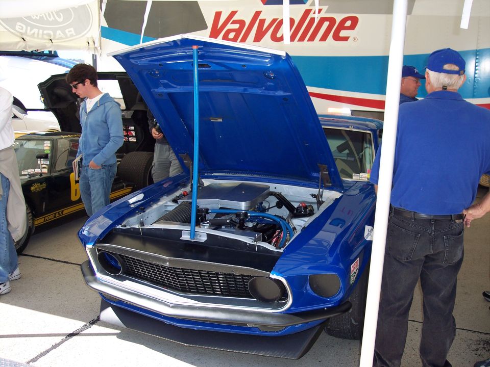 Blue 1969 Ford Mustang with hood open, showing engine bay. Another black car with yellow stripe is visible to the left