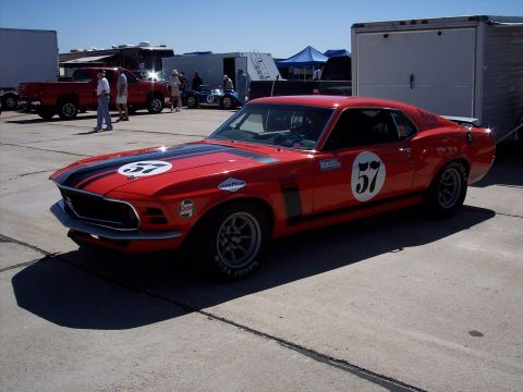 1971 Ford Mustang - A vintage Mustang race car is parked on asphalt with other vehicles and trailers in the background under a clear sky.