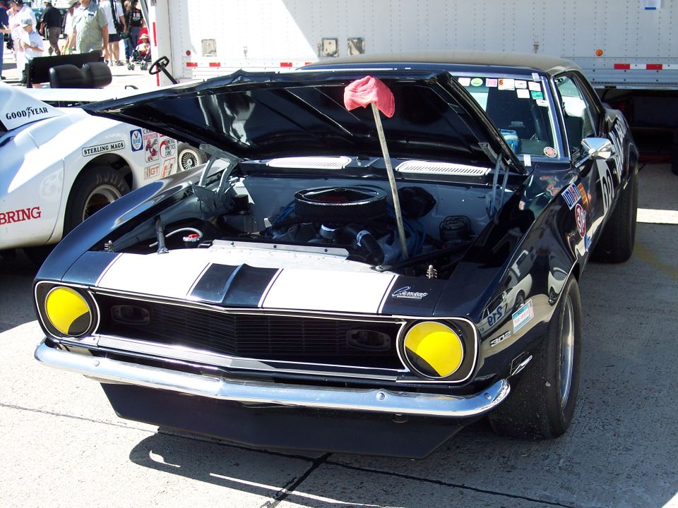 Black 1969 Chevrolet Camaro SS with white racing stripes and yellow headlights, hood open. Another white car is partially visible to the left