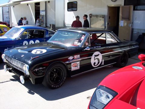 1971 Ford Mustang - A black Ford Falcon Sprint race car is parked at a track event, with other vehicles and people in the background.