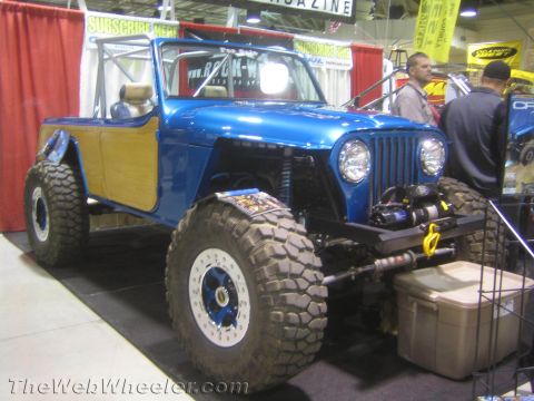 1990 Ford Bronco - blue custom open-top 4x4 with wood-grain side panels, front winch and large tires on beadlock wheels displayed at an indoor show.